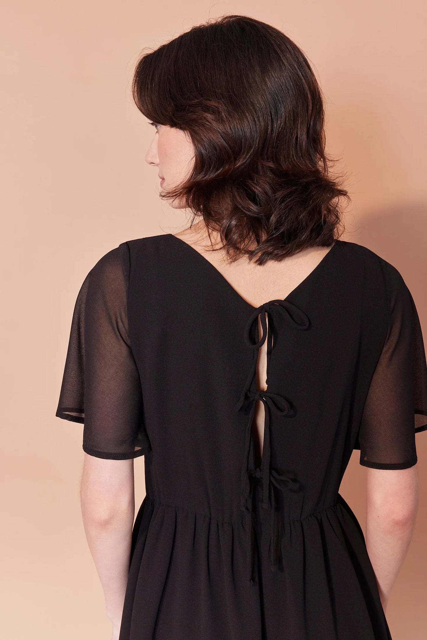 Back waist-up view of  woman wearing the Sisi Dress by Cherry Bobin in Black,  a chiffon dress with a v-neck front and back, short full sleeves, a gathered waist, an open back with tie closures, and an A-line skirt that falls above the knee. She is standing in front of a beige background. 