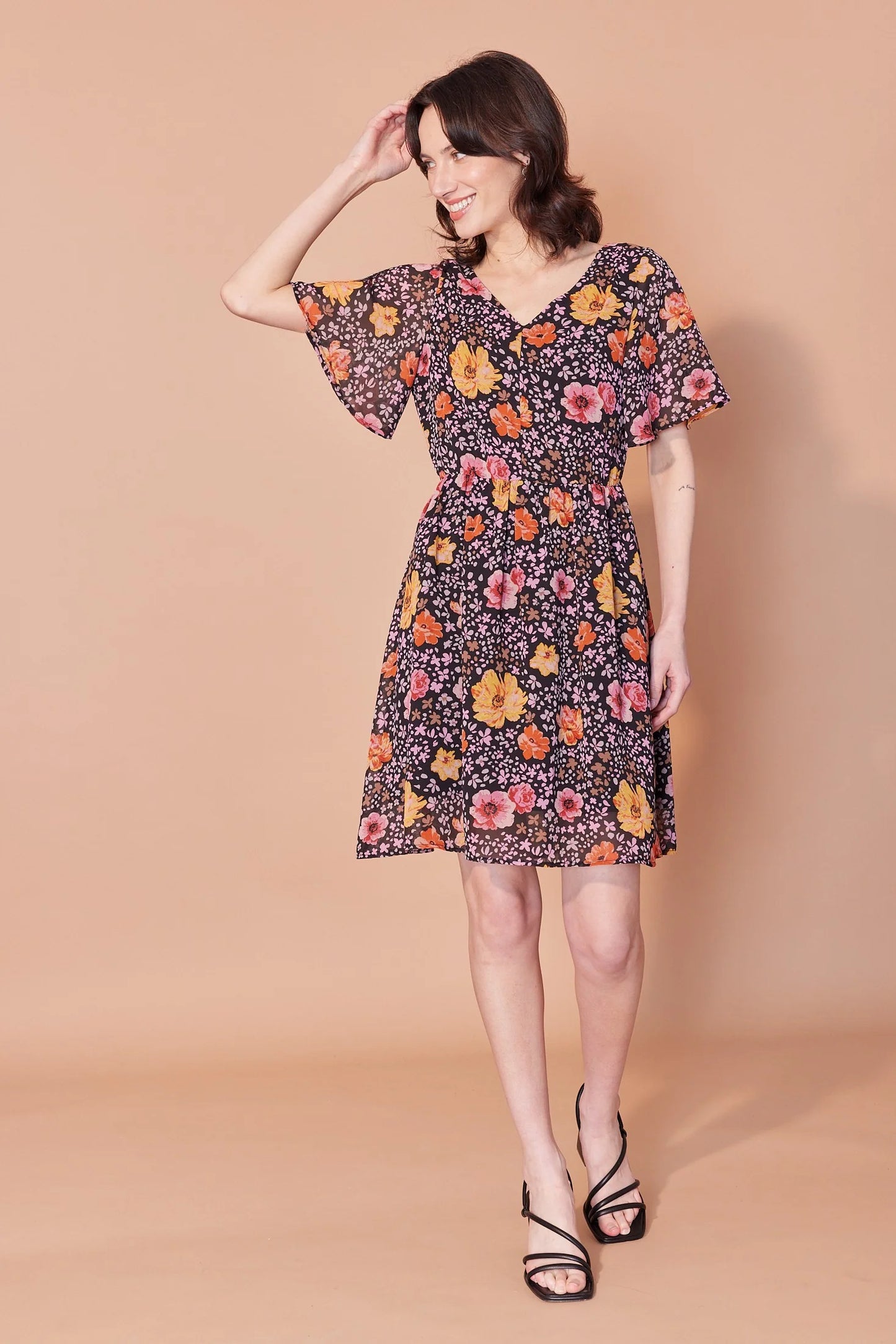 A woman wearing the Sisi Dress by Cherry Bobin in Flowers and Petals print, a chiffon dress with a v-neck front and back, short full sleeves, a gathered waist, an open back with tie closures, and an A-line skirt that falls above the knee. She is standing in front of a beige background. 