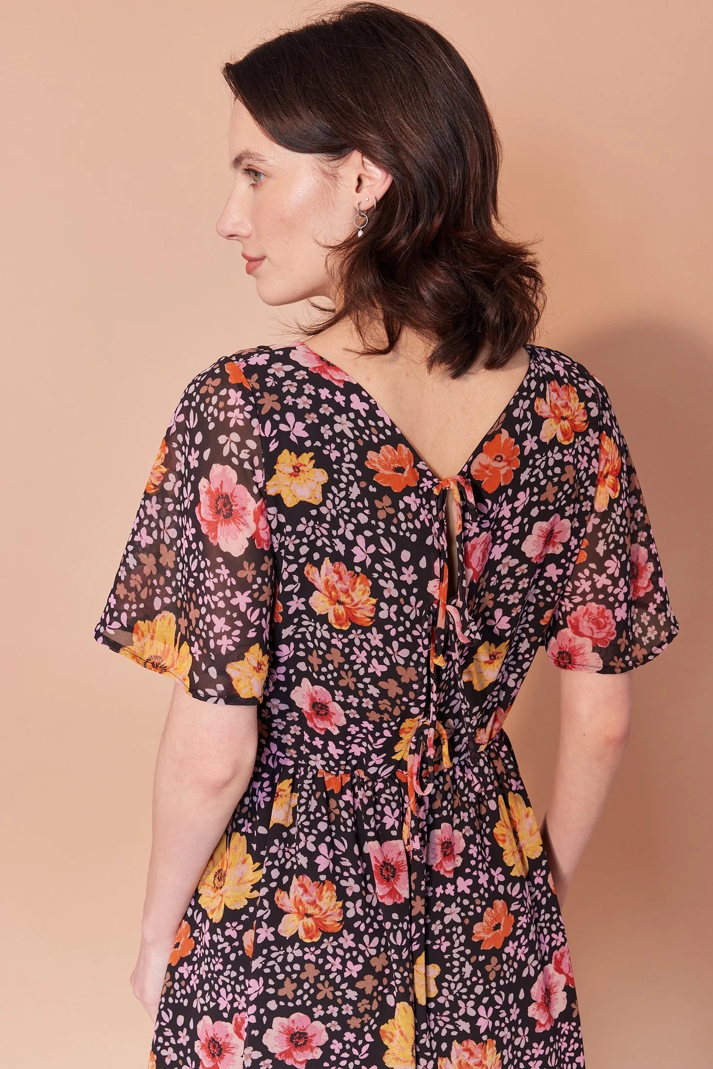 Back waist-up view of a woman wearing the Sisi Dress by Cherry Bobin in Flowers and Petals print, a chiffon dress with a v-neck front and back, short full sleeves, a gathered waist, an open back with tie closures, and an A-line skirt that falls above the knee. She is standing in front of a beige background. 