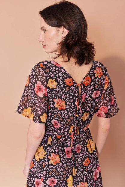 Back waist-up view of a woman wearing the Sisi Dress by Cherry Bobin in Flowers and Petals print, a chiffon dress with a v-neck front and back, short full sleeves, a gathered waist, an open back with tie closures, and an A-line skirt that falls above the knee. She is standing in front of a beige background. 