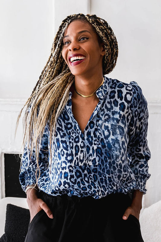 Waist-up shot of a woman wearing the Blue Leopard Blouse by Dotty, featuring a V-neck, banded collar, and long sleeves with smocked cuffs. It has a flowy fit and and rounded hem. She is wearing it with black pants and standing in front of a white wall. 