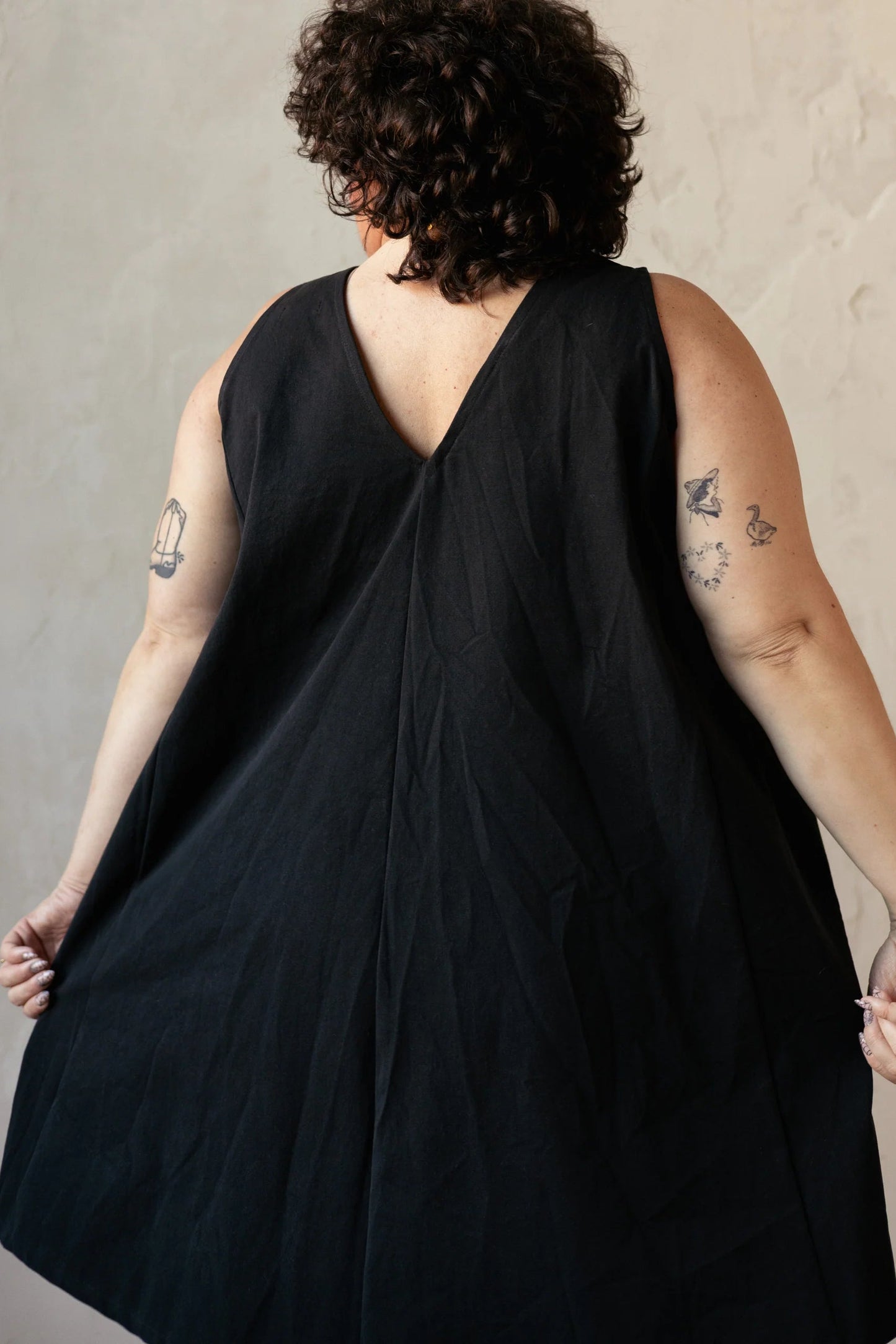 Back view of a woman wearing the Caro Dress by Lights of All in Black, a sleeveless knee-length trapeze dress with a round neck at the front and a V at the back. She is standing in front of a beige background. 