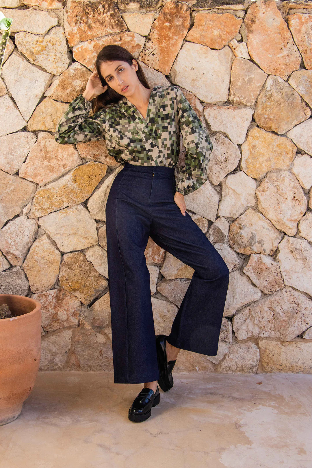 A woman wearing the Manama Blouse by MAS in a pixellated Camo Print, featuring a modest cross-over neckline, darts, and long sleeves with elastic cuffs, with the Vienna Pants in Jeans. She is standing in front of a stone wall. 
