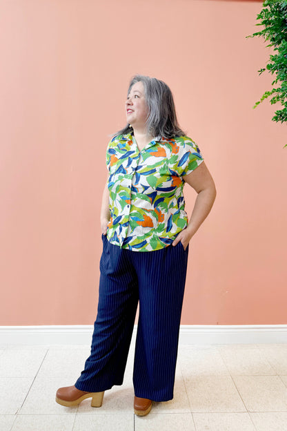 Christina is wearing the Eva Top by Pure Essence in Blue/Cilantro print, with a blouse collar, button front, short sleeves, and rounded hem. She is wearing it with the Eloise Pants in Ink pinstripe and standing in front of a coral wall. 