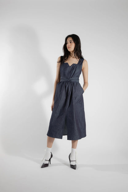 A woman wearing the Cornifle Dress by Eve Lavoie in Denim, a sleeveless dress with a fitted bodice, wide straps, square neckline with a V-notch, full skirt that falls below the knee, a front slit, and patch pockets. She is standing in front of a white background.  