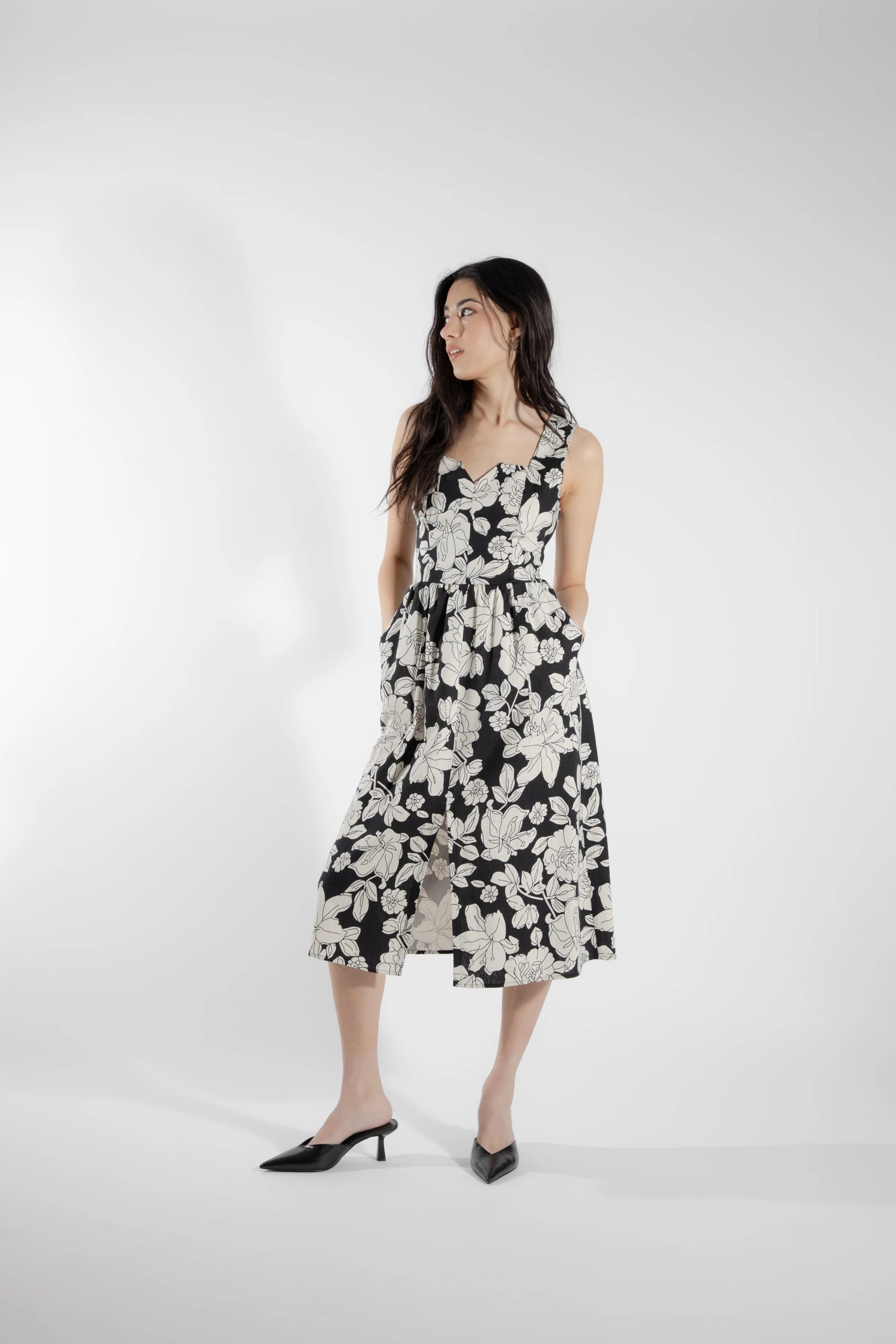 A woman wearing the Cornifle Dress by Eve Lavoie in black and white Floral, a sleeveless dress with a fitted bodice, wide straps, square neckline with a V-notch, full skirt that falls below the knee, a front slit, and patch pockets. She is standing in front of a white background.  