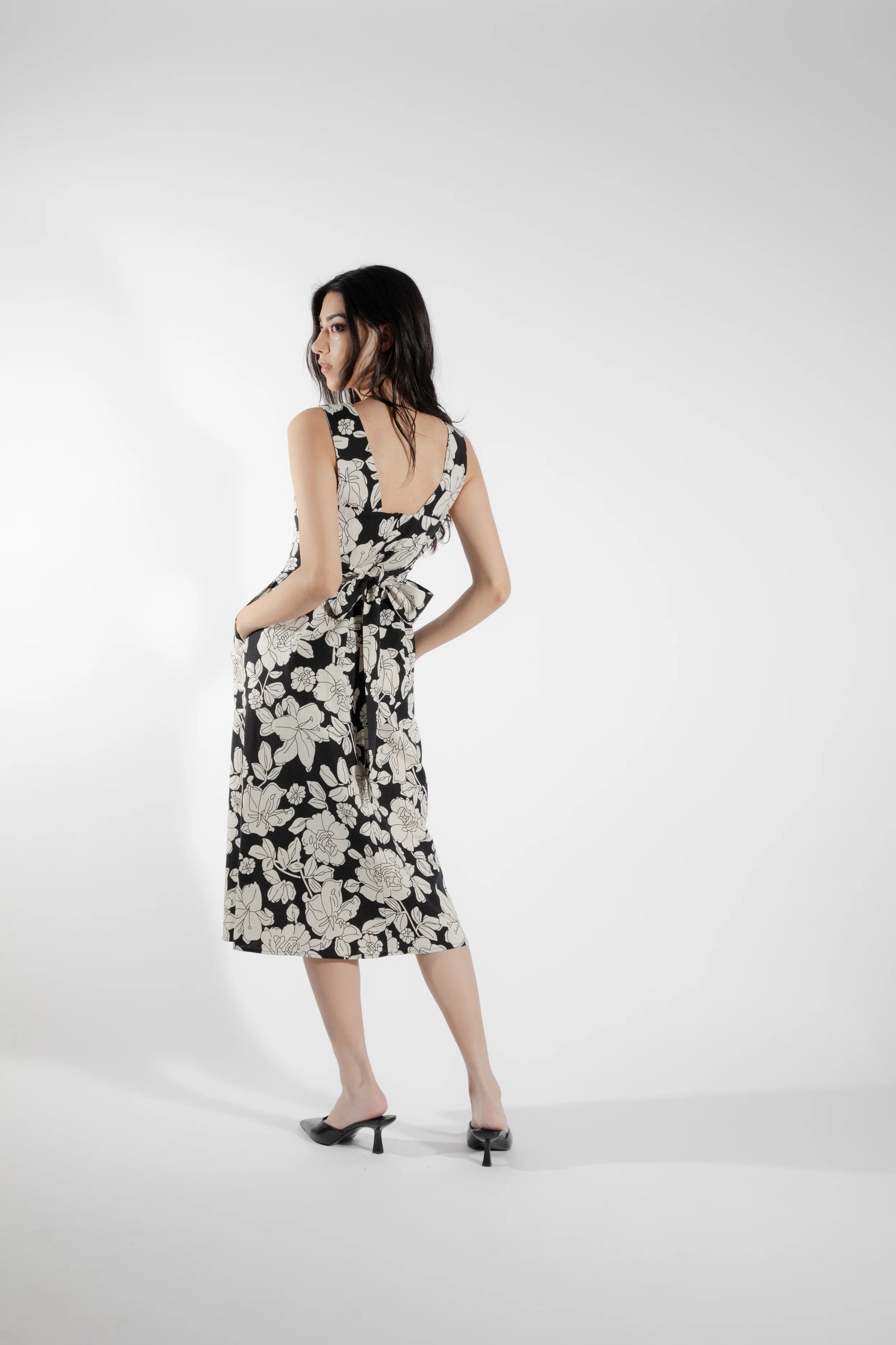 Back view of a woman wearing the Cornifle Dress by Eve Lavoie in black and white Floral, a sleeveless dress with a fitted bodice, wide straps, square neckline with a V-notch, full skirt that falls below the knee, a front slit, and patch pockets. She is standing in front of a white background.  