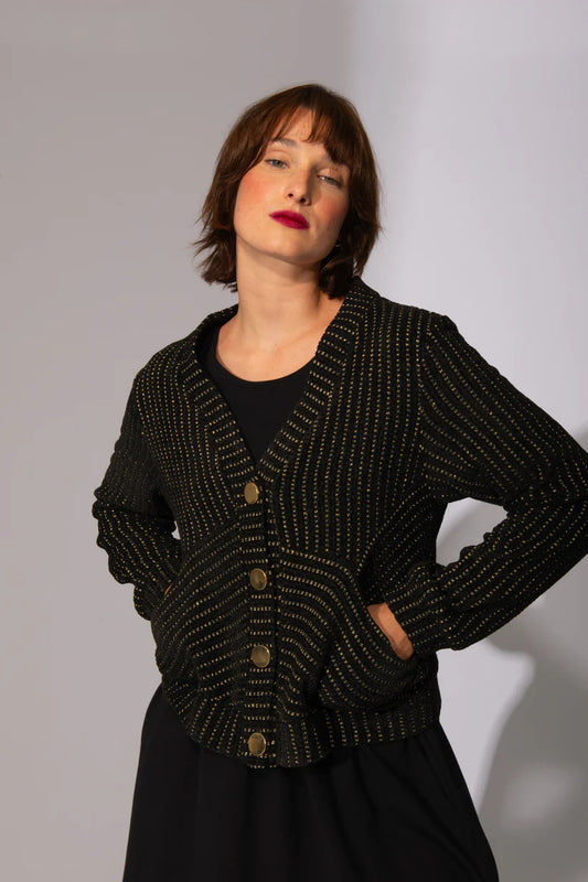 A woman wearing the Gold Lion Knit Cardigan by Eve Lavoie in Black, featuring a V-neck, gold thread embedded in the black fabric, gold buttons, and bias pockets. She is wearing it over a black dress and standing in front of a white background. 