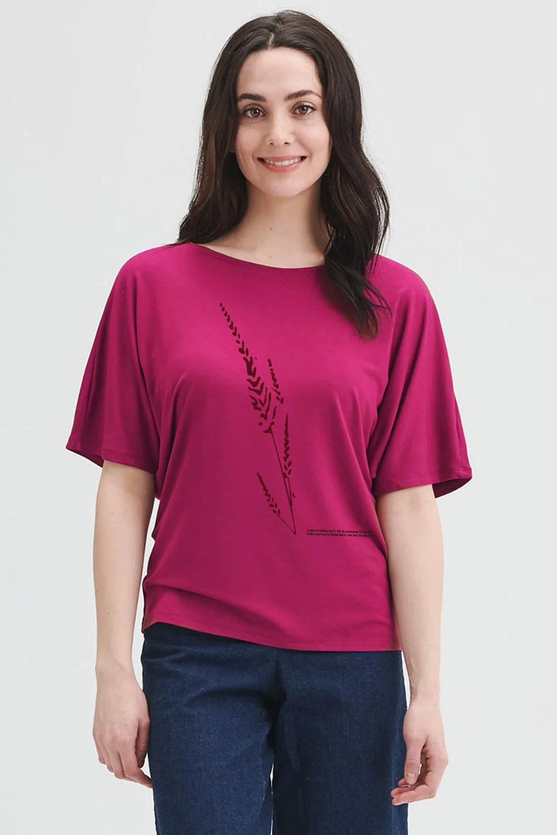 A woman wearing the Betty Top by Message Factory in Raspberry, a bamboo top with short extended sleeves, a plant print, and French inscription about nature. She is wearing it with jeans and standing in front of a white background. 
The quote reads: La nature a sa facon  bien a elle de nous ramener a l'essentiel. Souple comme un brin d'herbe dans le vent, tout devient possible. 