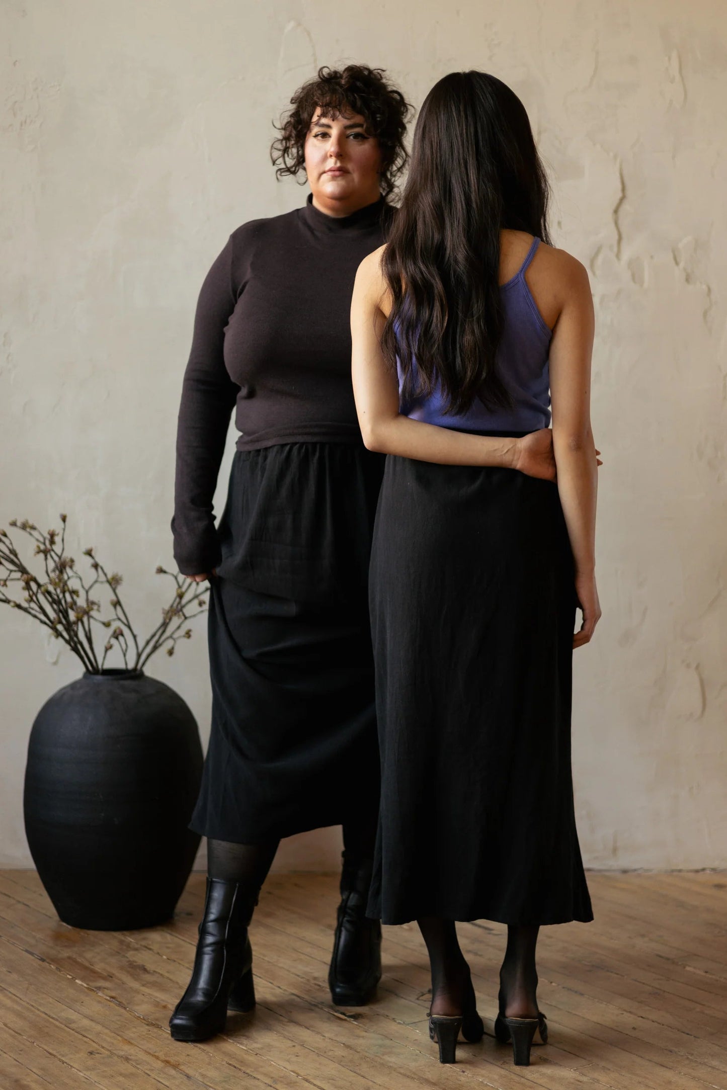 Two woman wearing the Simone Skirt by Lights of All in Black, a sleek calf-length skirt with a flared hem and an elastic waist. One is facing backwards and is wearing it with a purple top, the other is facing forwards and is wearing it with a brown turtleneck. They are standing in a room with furniture. 
