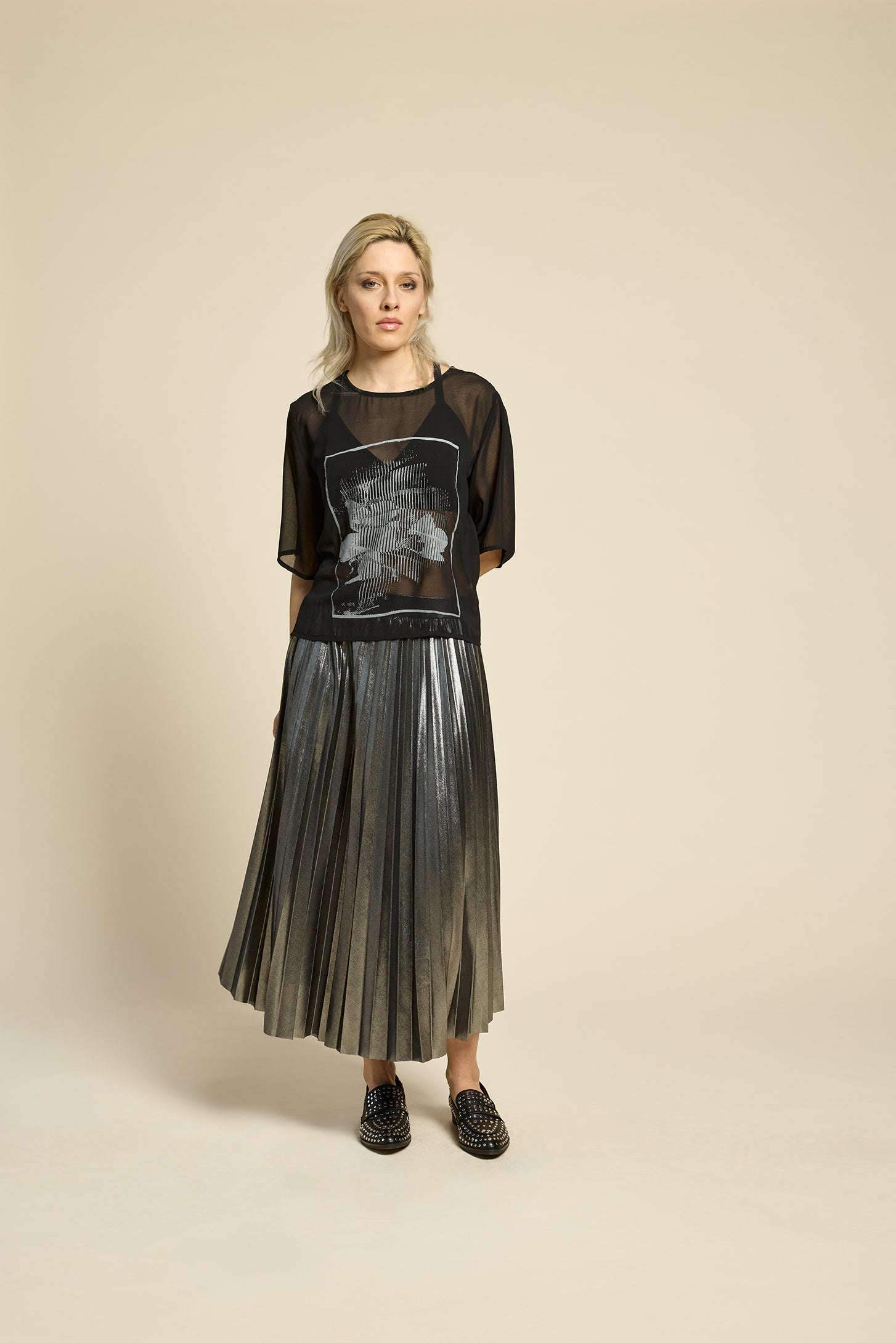 A woman wearing the Feyer Skirt by Cokluch in Glow Knit Silver, a midi length pleated skirt in shimmery metallic fabric. She is wearing it with a sheer black graphic top and is standing in front of a beige background. 