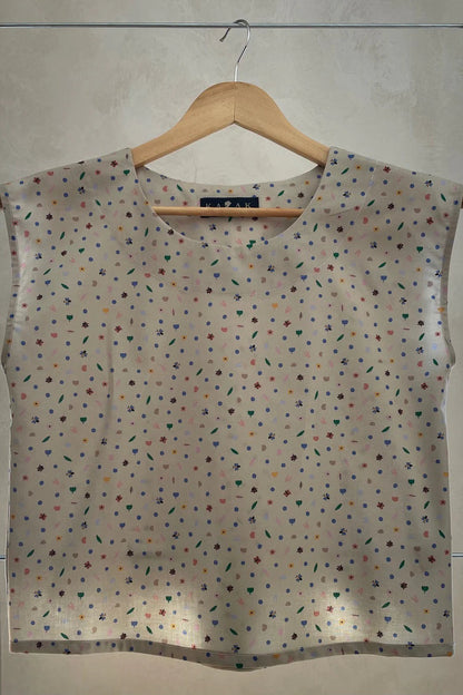 The Sandrigo Top from Kazak in Flowers and Insects print, a cotton top with short extended sleeves and a wide round neck, is shown on a wooden hanger against a grey background. 