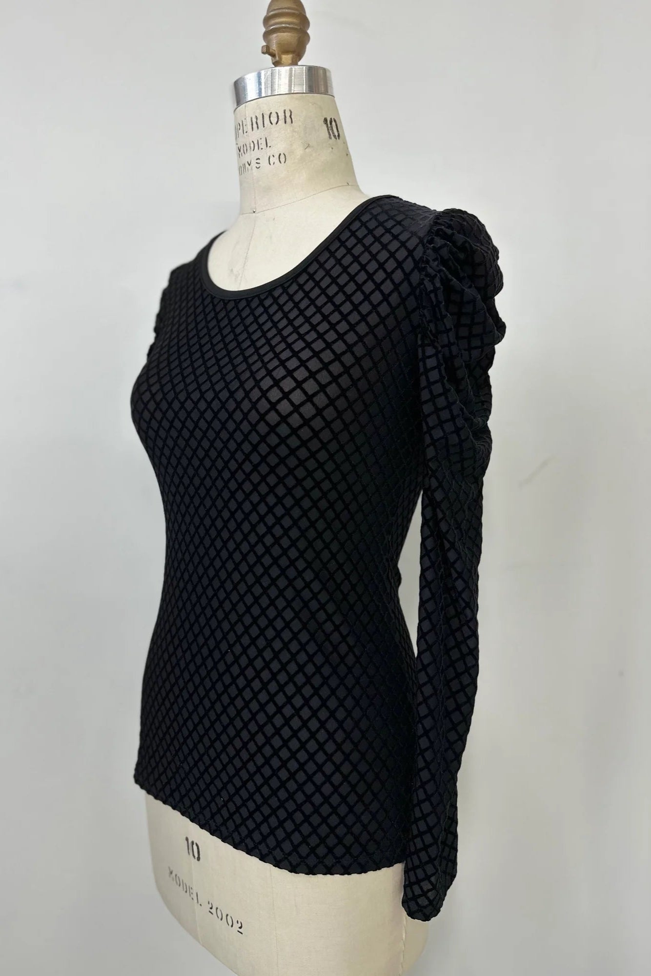 The Octavia Sweater by Kollontai, featuring black cut velvet in a diamond pattern, gathered shoulders, long sleeves, an oval neckline, and a slim fit, is shown on mannequin against a white background. 