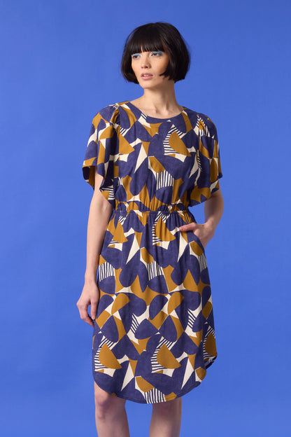 3/4 shot of a woman wearing the Gemma Dress by Cokluch in a Cumin half-moon print, standing in front of a blue background 