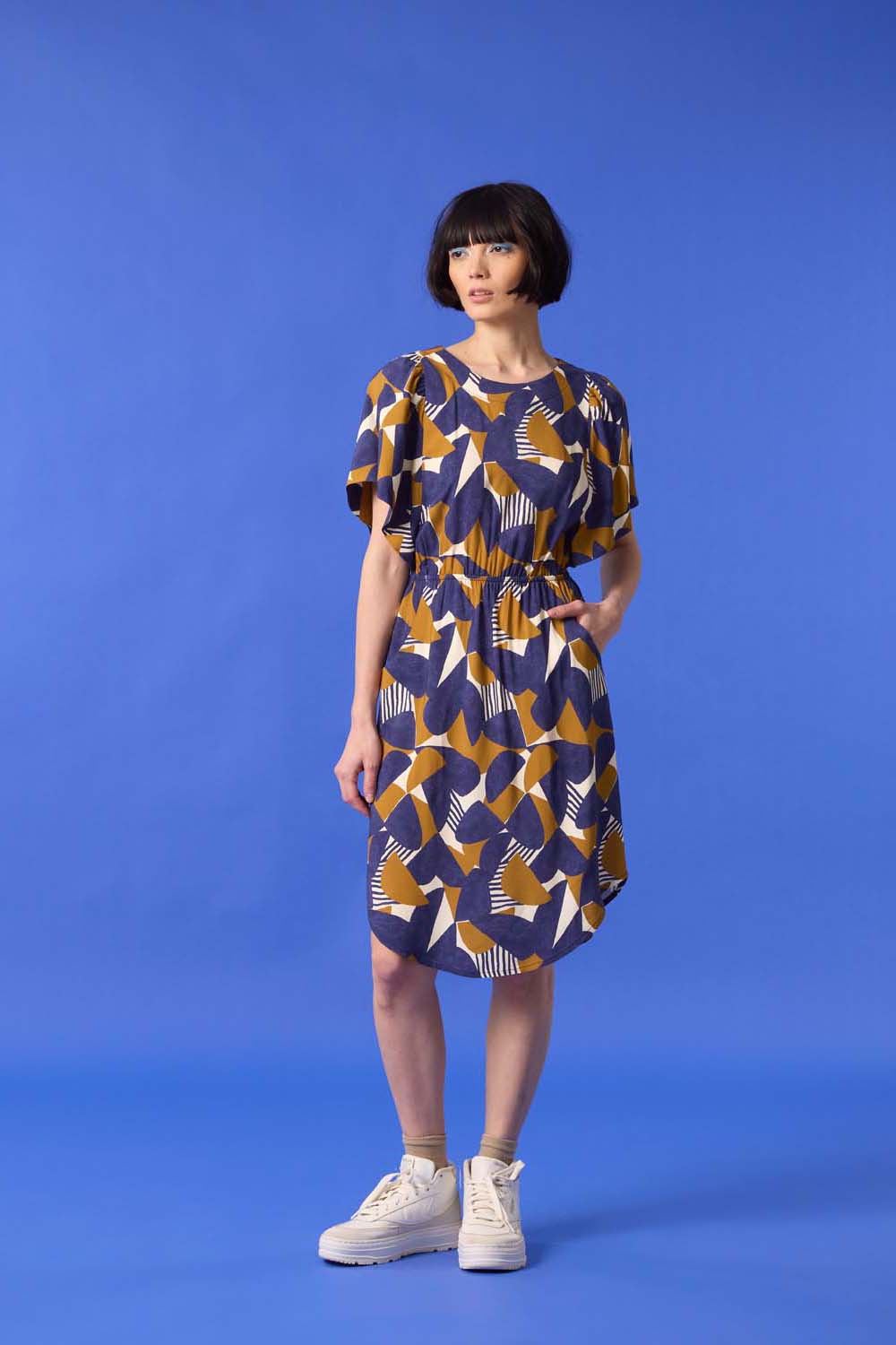 A woman wearing the Gemma Dress by Cokluch in a Cumin half-moon print, standing in front of a blue background 