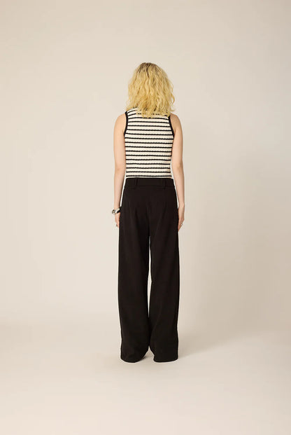 Back view of a woman wearing the Hebe Shirt from Cokluch, a black and white striped sleeveless top with a round neck. She is wearing it with black pants against a beige background