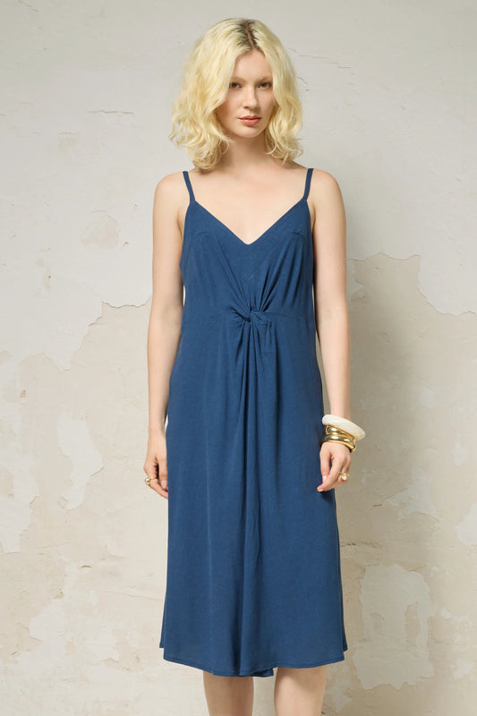 A woman wearing the Hera Dress by Cokluch in Denim, a spaghetti strap midi dress with a V-neck and a knot detail at the front waist. She is standing against a beige wall. 