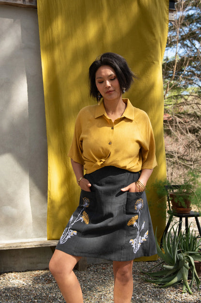 A woman wearing the Vicenze Screen-printed Skirt by Kazak in Black with Dandelions, an above the knee A-line skirt with hand-screened flowers, an elasticated waist, and large patch pockets. She is wearing it with the  Hosta shirt in Mustard and standing outside by a grey wall and white and yellow curtains.