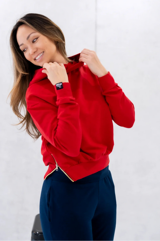 3/4 view of a woman wearing the Olive+Splash Bamboo Bomber Hoodie in Red, a cropped hoodie with long sleeves and a vertical gold zipper detail on the right hem. She is wearing it with Navy Pants and standing in front of a white background. 