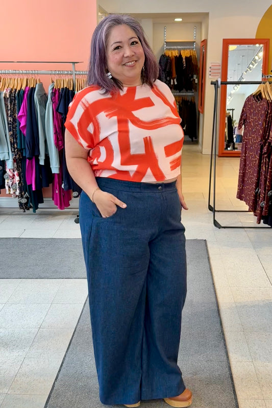 A woman wearing the Nyx top from Cockluch in Red Shodo, a short sleeved, loose fitting top with a drawstring detail at the bottom hem. She is wearing it with the atisse Trousers  by Melow and standing inside Flock. 