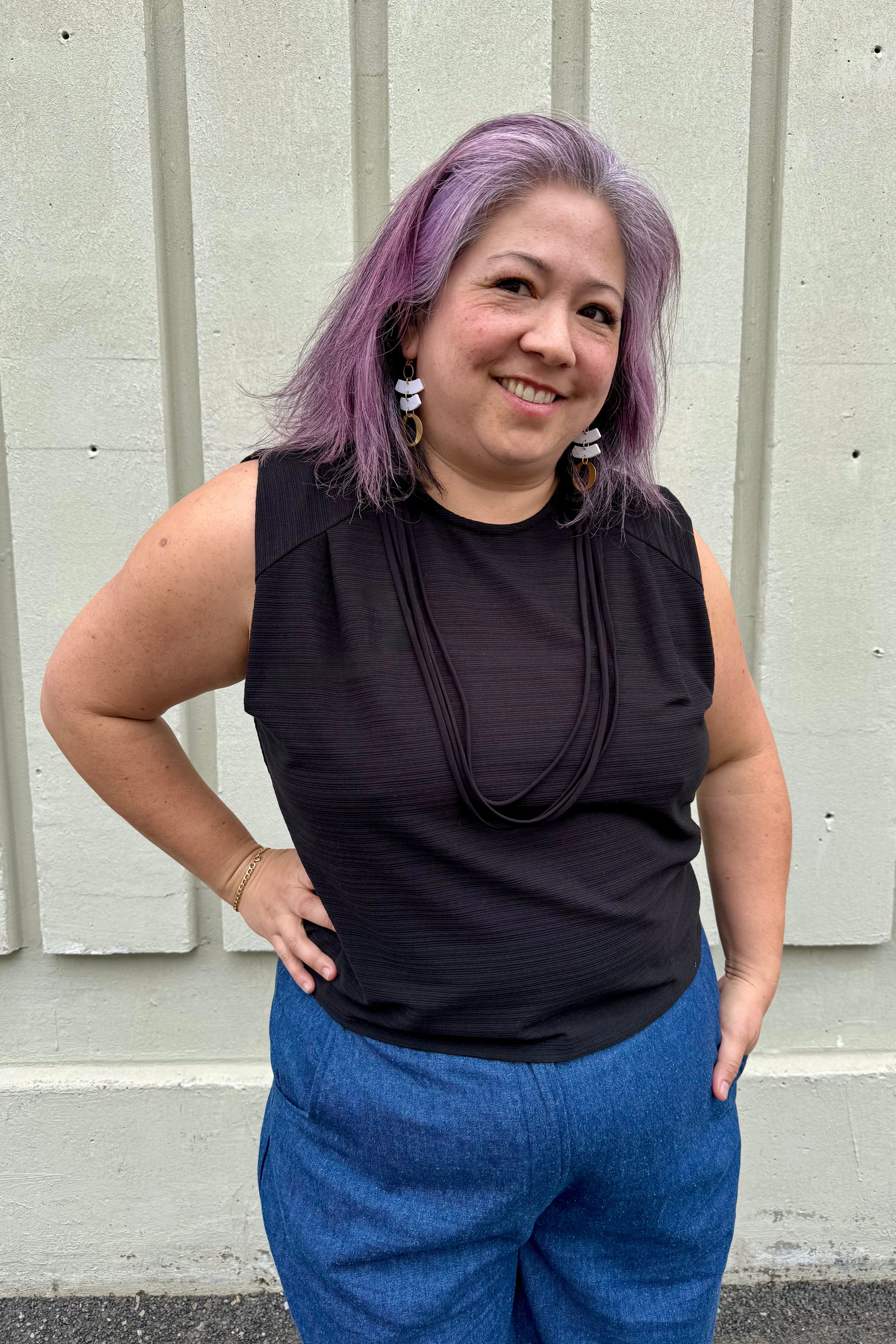 A woman wearing the Path Top from Cokluch in Black, a sleeveless top with line details at the shoulders and decorative straps that attach at each shoulder and hang to the front. She is wearing it with the Matisse pants by Melow and standing outside in front of a panelled wall. 