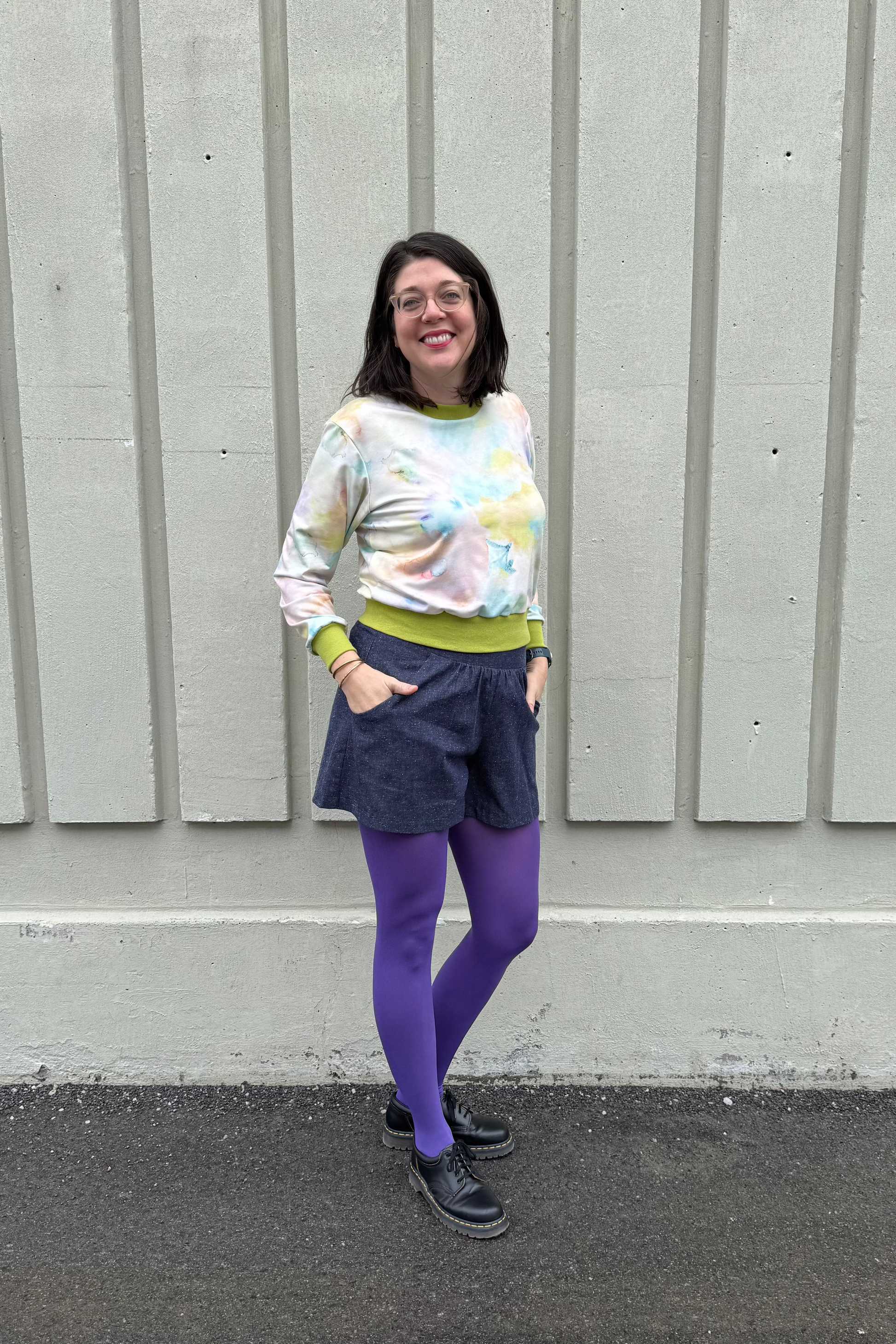 A woman wearing the Maya shorts from Cokluch in Indigo Speckle, lightweight denim shorts with a waist that's elasticized at the back, wide culotte style legs, and a side pockets. She is wearing them with the Wanda E26 Sweater by Slak in Watercolour print and purple tights and standing outside in front of panelled wall. 