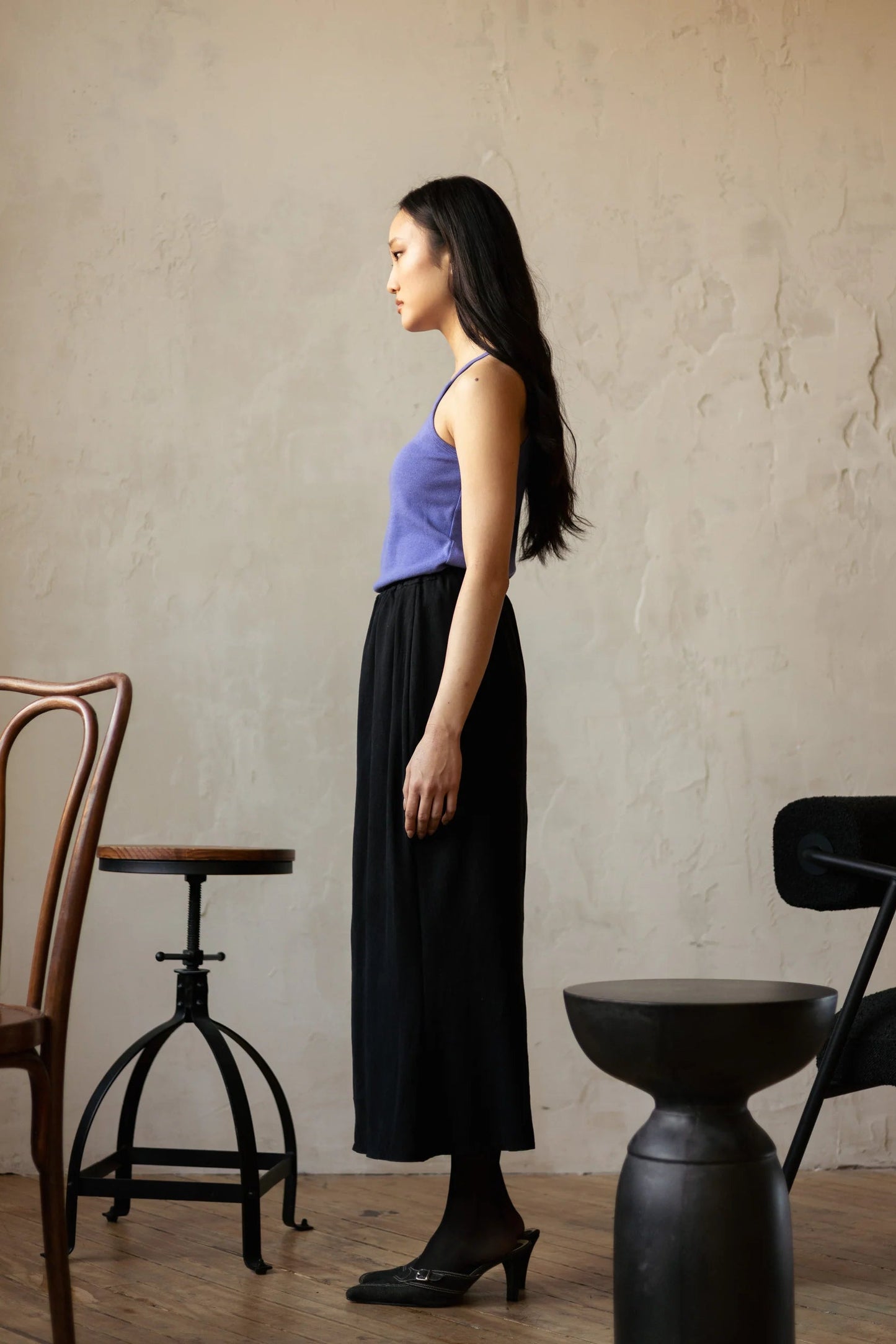 Side view of a woman wearing the Simone Skirt by Lights of All in Black, a sleek calf-length skirt with a flared hem and an elastic waist. She is wearing it with a purple top and standing in a room with furniture. 