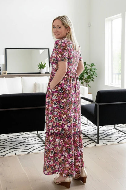 Back view of a woman wearing the Petal Print by Dotty in Pink/Blue floral,  a maxi dress with short flutter sleeves, a split V-neck and ruffled collar, with a gathered waist and A-line skirt. She is standing in a modern living room.
