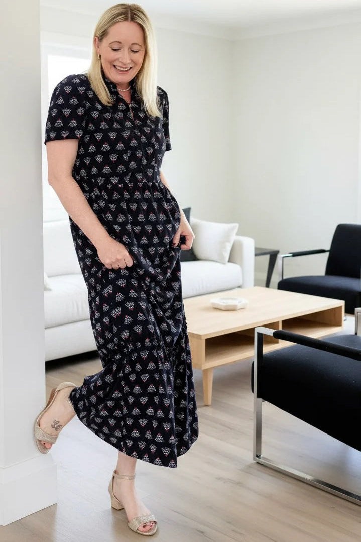 A woman wearing the Art Deco Dress by Dotty in Black print, a maxi dress with short flutter sleeves, a split V-neck and ruffled collar, with a gathered waist and A-line skirt. She is standing in a modern living room.