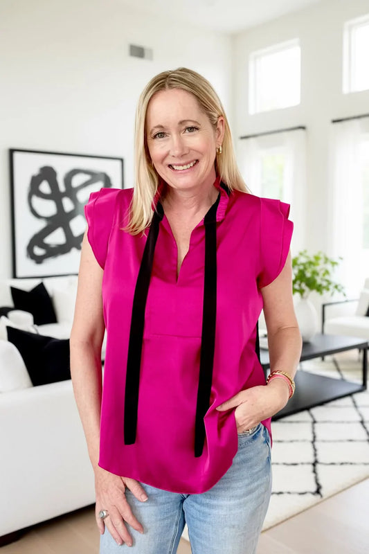 A woman wearing the Satin Tie blouse by Dotty in Pink, a short fluttery sleeved blouse with black tie at the split V-neck. She is wearing it with light blue jeans, standing in a bright modern living room.