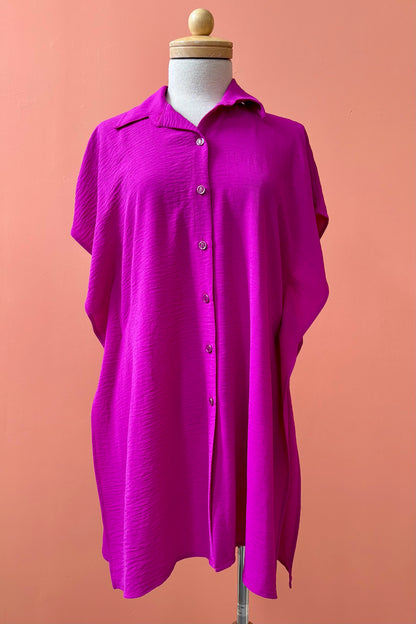 The Geneva Top by Pure Essence in Magenta, a long and loose button front shirt with short sleeves, a classic collar, and high side slits is shown on a mannequin against a coral background. 