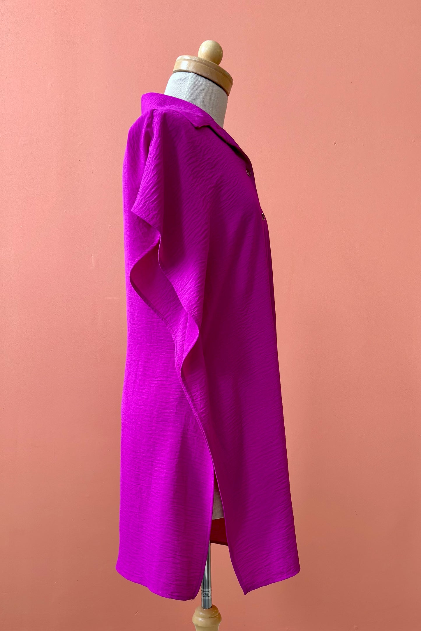 A side view of the Geneva Top by Pure Essence in Magenta, a long and loose button front shirt with short sleeves, a classic collar, and high side slits is shown on a mannequin against a coral background. 