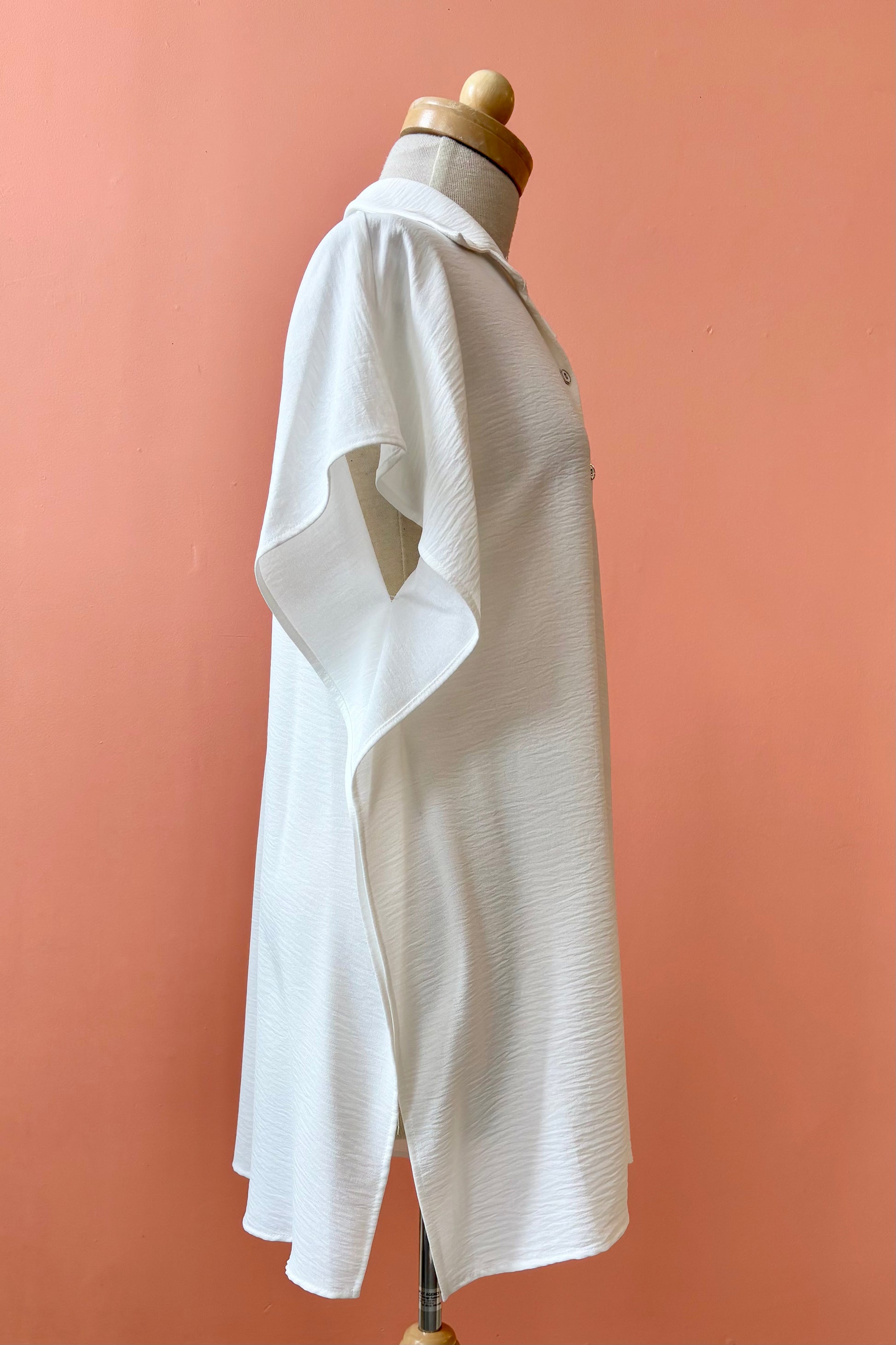 The Geneva Top by Pure Essence in White, a long and loose button front shirt with short sleeves, a classic collar, and high side slits is shown on a mannequin against a coral background. 