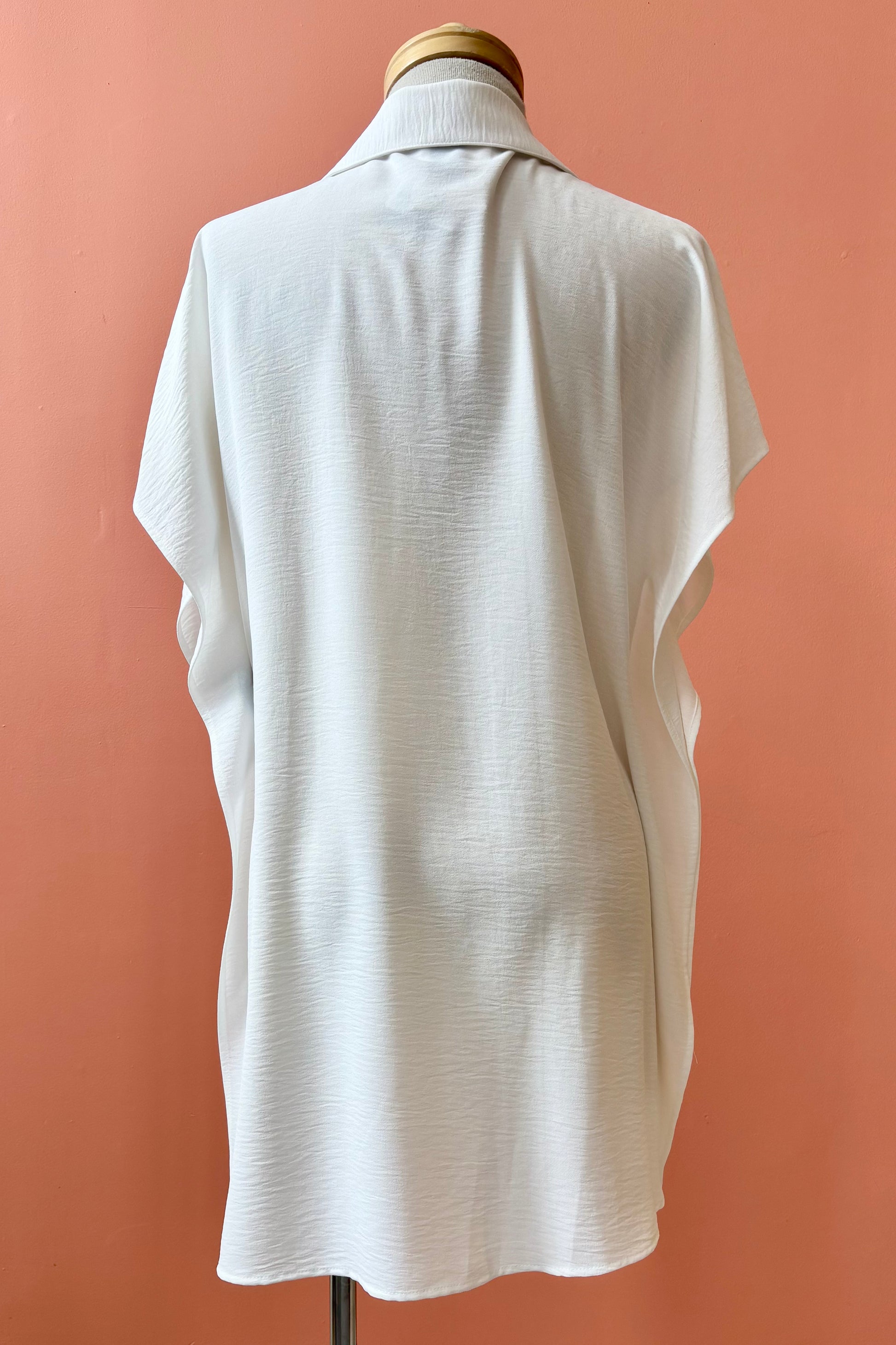 A back view of the Geneva Top by Pure Essence in White, a long and loose button front shirt with short sleeves, a classic collar, and high side slits is shown on a mannequin against a coral background. 