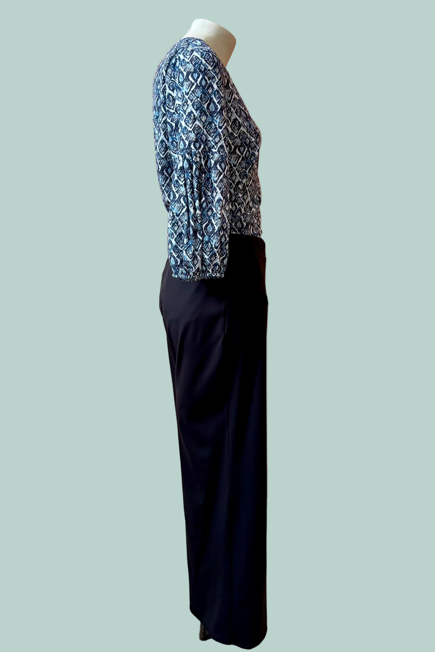 Side view of a mannequin wearing a the Gloria Pants by Pure Essence in Black with a blue patterned top on a light teal background