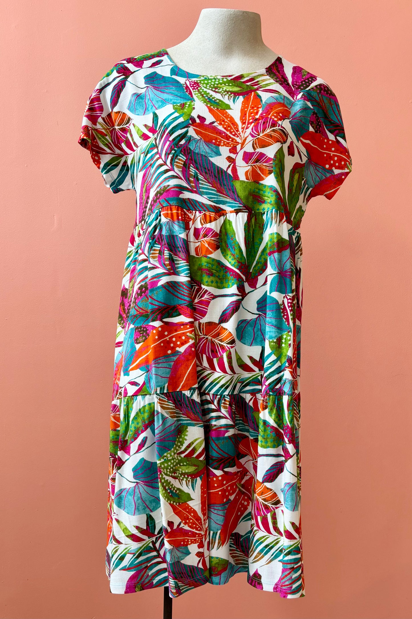 The Grayson Dress by Pure Essence in Peacock Palms, a knee-length baby doll style dress with a tiered skirt and short sleeves, is shown on a mannequin against a coral  wall. 