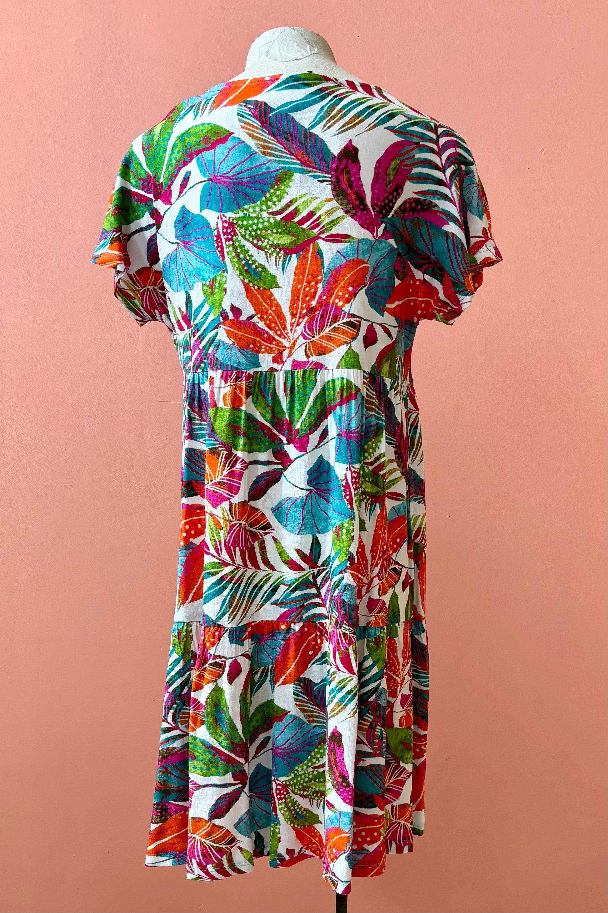 Back view of the Grayson Dress by Pure Essence in Peacock Palms, a knee-length baby doll style dress with a tiered skirt and short sleeves, is shown on a mannequin against a coral  wall. 
