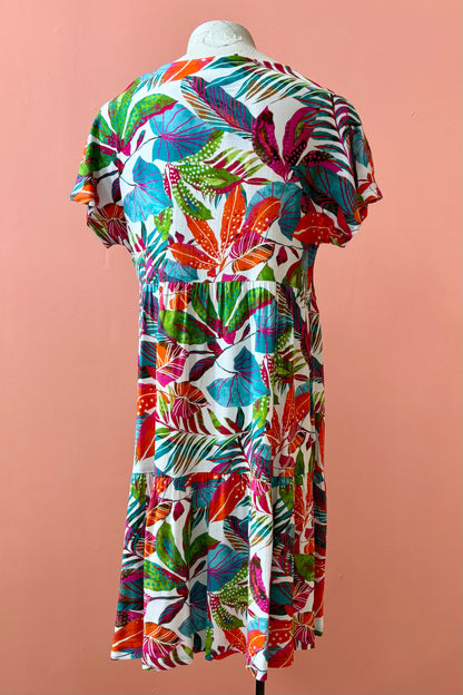 Back view of the Grayson Dress by Pure Essence in Peacock Palms, a knee-length baby doll style dress with a tiered skirt and short sleeves, is shown on a mannequin against a coral  wall. 