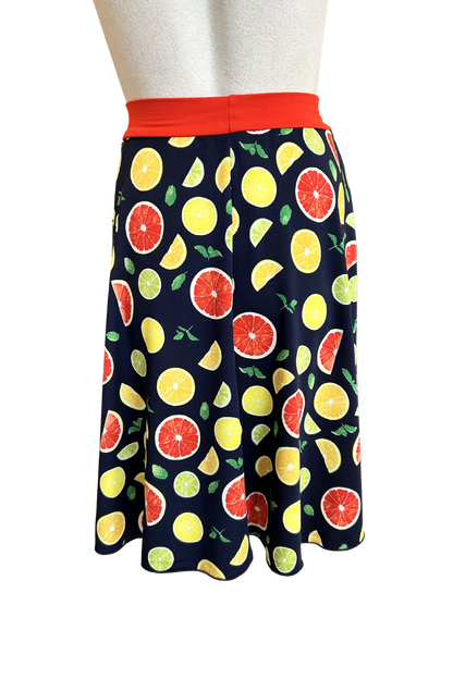 A back view of the Flirt Skirt by Slak in Citrus Print, an above the knee A-line skirt with a contrasting pull-on waistband, is shown on a mannequin against a white background. 