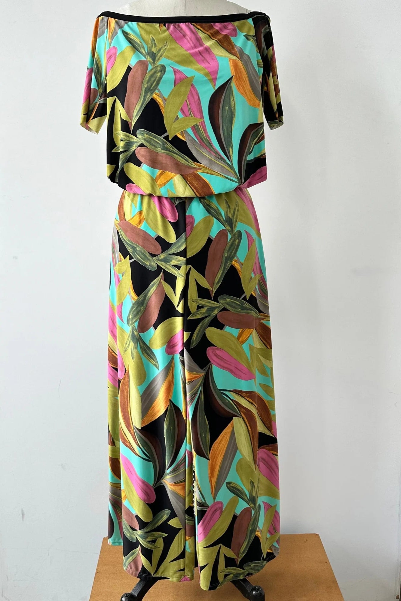  The Anthurium Jumpsuit by Kollontai in a Multicolour tropical print is show on a mannequin in front of a white background. It has an off the shoulder neckline, bloused elastic waist, short sleeves and wide 7/8 length legs.