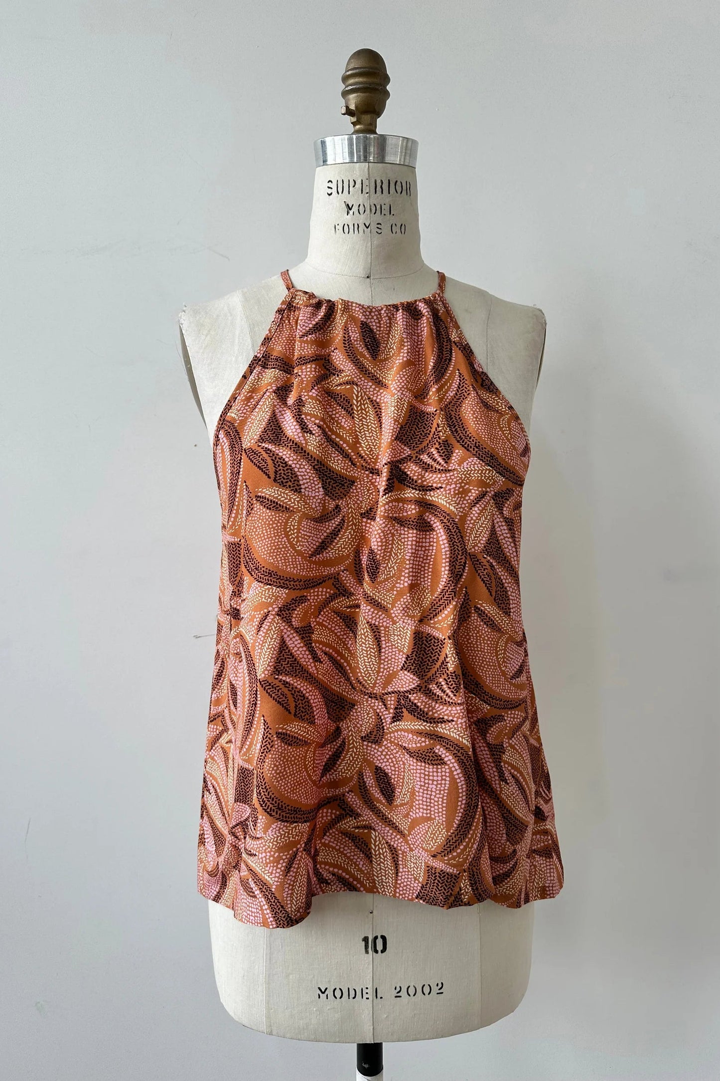 The Semira Camisole by Kollontai in Cinnamon print, a lightweight top with an adjustable halter neck and a loose fit, is shown on a mannequin in front of a white background. 