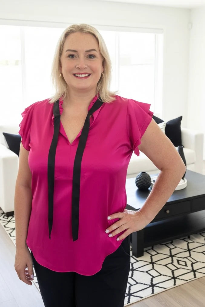 A woman wearing the Satin Tie blouse by Dotty in Pink, a short fluttery sleeved blouse with black tie at the split V-neck. She is wearing it with black pants and standing in a bright modern living room.