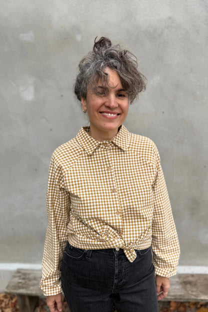 A woman wearing the Acer Blouse by Kazak in Ochre Gingham, a raglan sleeved blouse with a classic collar, button front, and elastic cuffs. She is wearing it with black jeans and standing in front of a grey wall. 