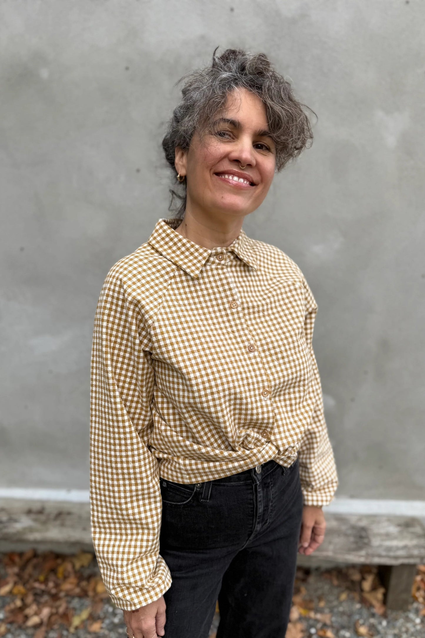 A woman wearing the Acer Blouse by Kazak in Ochre Gingham, a raglan sleeved blouse with a classic collar, button front, and elastic cuffs. She is wearing it with black jeans and standing in front of a grey wall. 