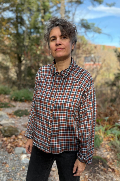 A woman wearing the Acer Blouse by Kazak in Checkered fabric, a raglan sleeved blouse with a classic collar, button front, and elastic cuffs. She is wearing it with black jeans and standing in a forest.