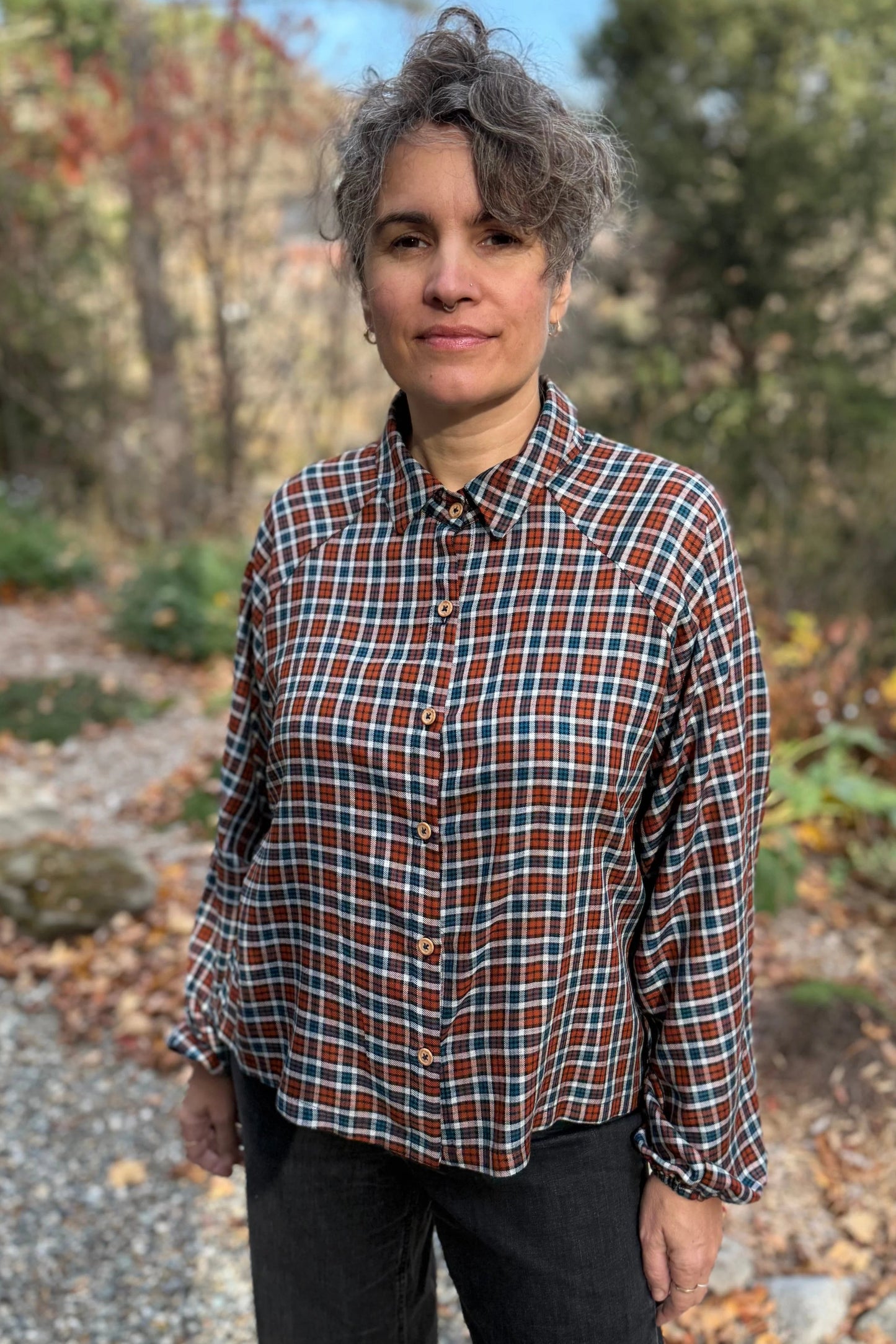 A woman wearing the Acer Blouse by Kazak in Checkered fabric, a raglan sleeved blouse with a classic collar, button front, and elastic cuffs. She is wearing it with black jeans and standing in a forest.