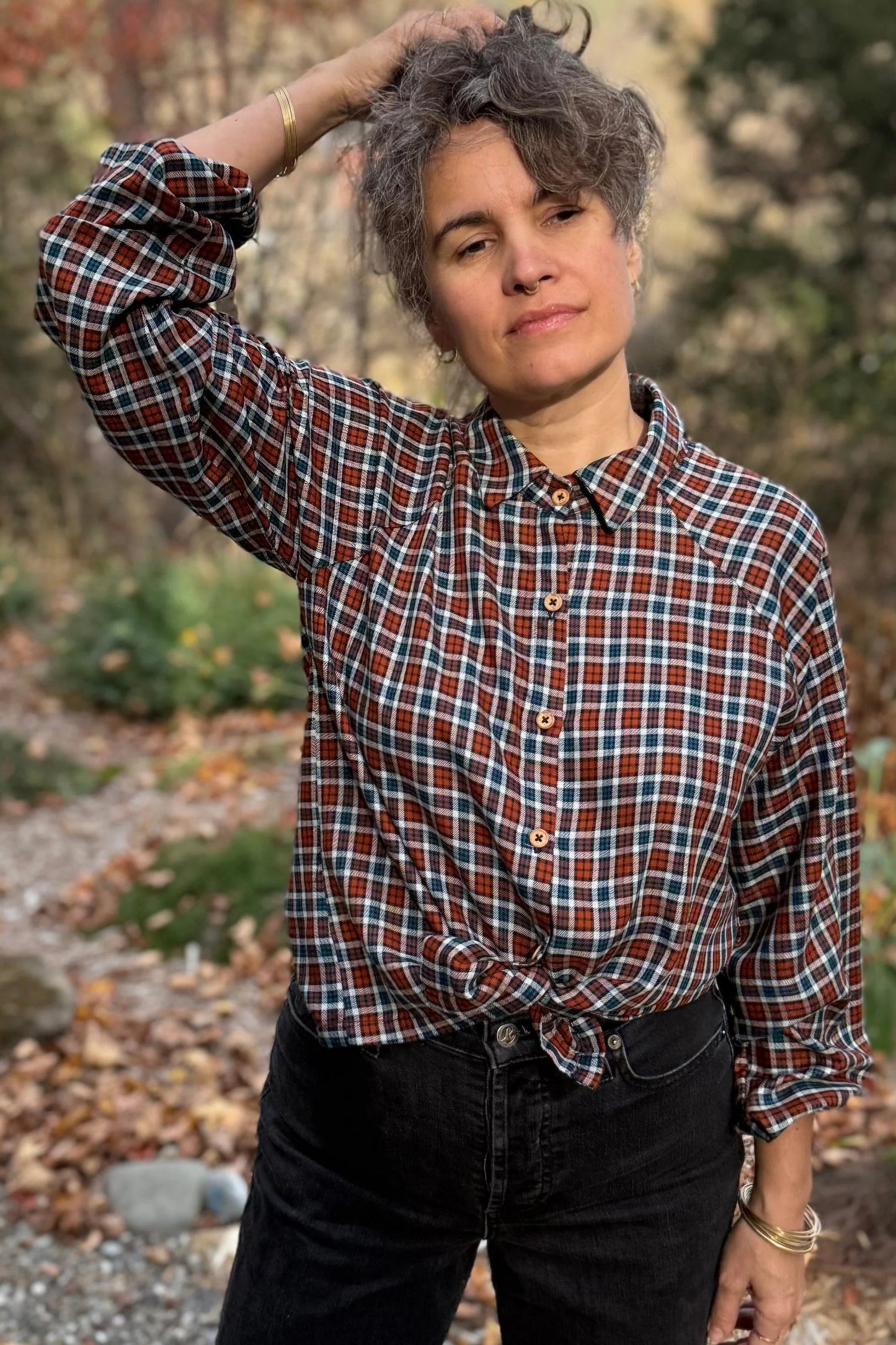 A woman wearing the Acer Blouse by Kazak in Checkered fabric, a raglan sleeved blouse with a classic collar, button front, and elastic cuffs. She is wearing it with black jeans and standing in a forest.