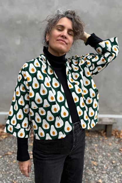 A woman wearing the Magnolia Jacket by Kazak in green and white corduroy Pears print fabric, a short kimono style open jacket with 3/4 sleeves and large patch pockets. She is wearing it with a black top and black jeans and standing in front of a grey wall. 