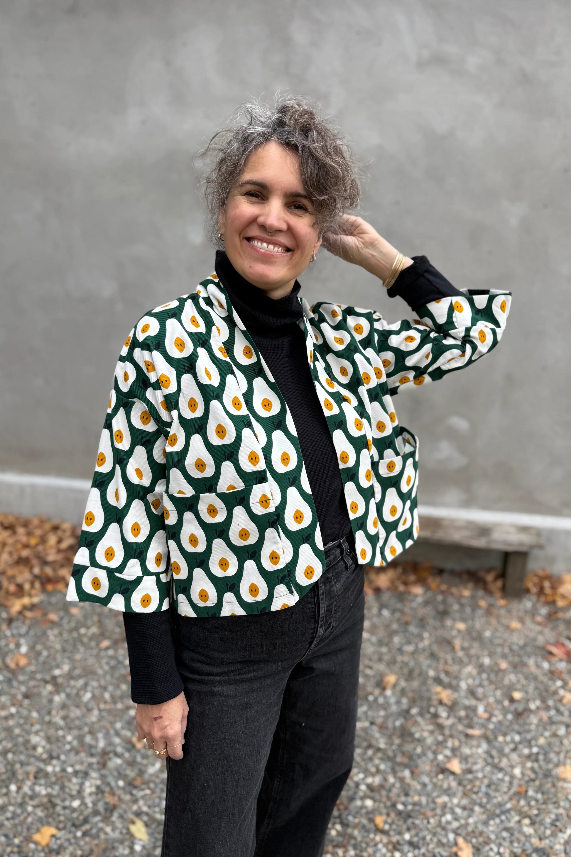 A woman wearing the Magnolia Jacket by Kazak in green and white corduroy Pears print fabric, a short kimono style open jacket with 3/4 sleeves and large patch pockets. She is wearing it with a black top and black jeans and standing in front of a grey wall. 