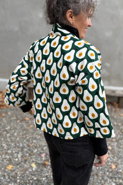 Back view of a woman wearing the Magnolia Jacket by Kazak in green and white corduroy Pears print fabric, a short kimono style open jacket with 3/4 sleeves and large patch pockets. She is wearing it with a black top and black jeans and standing in front of a grey wall. 